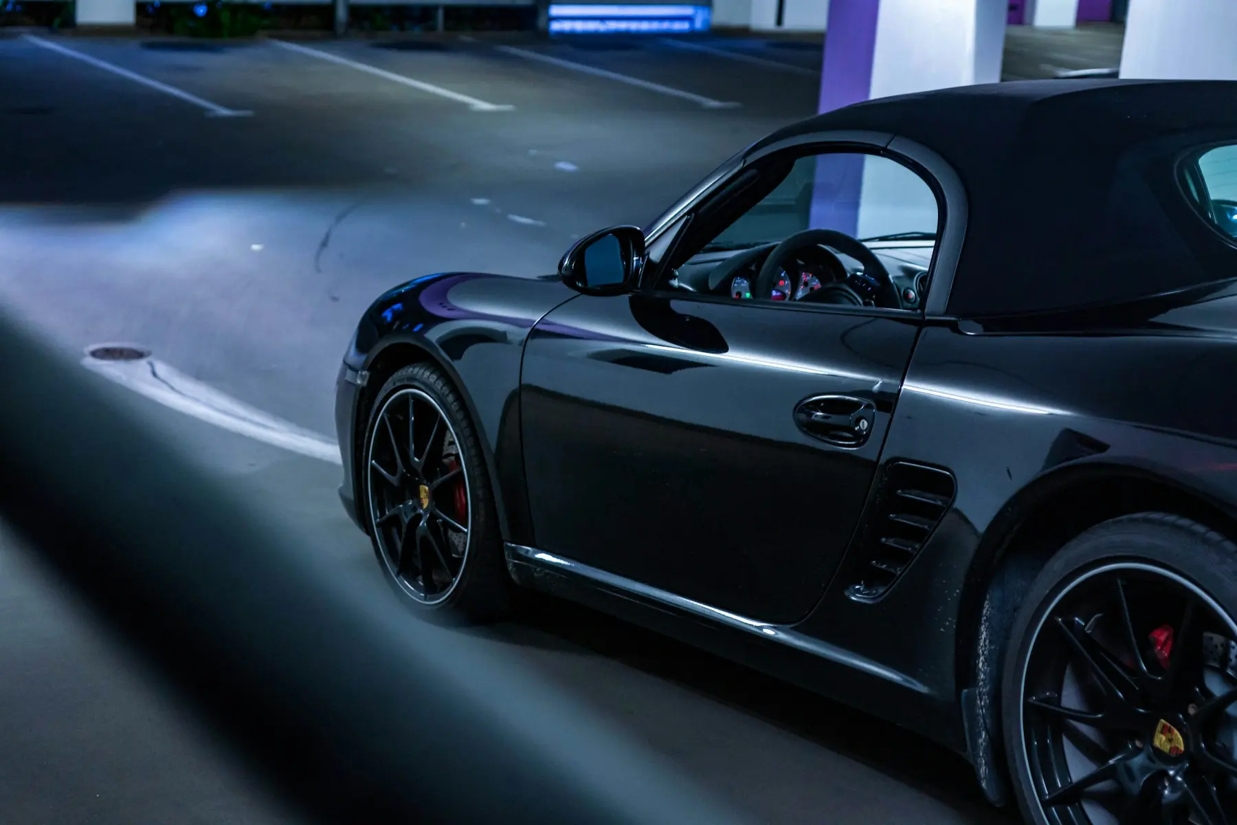 An angled side view of a dark Porsche convertible, its curved bodywork catching light inside a concrete parking structure.