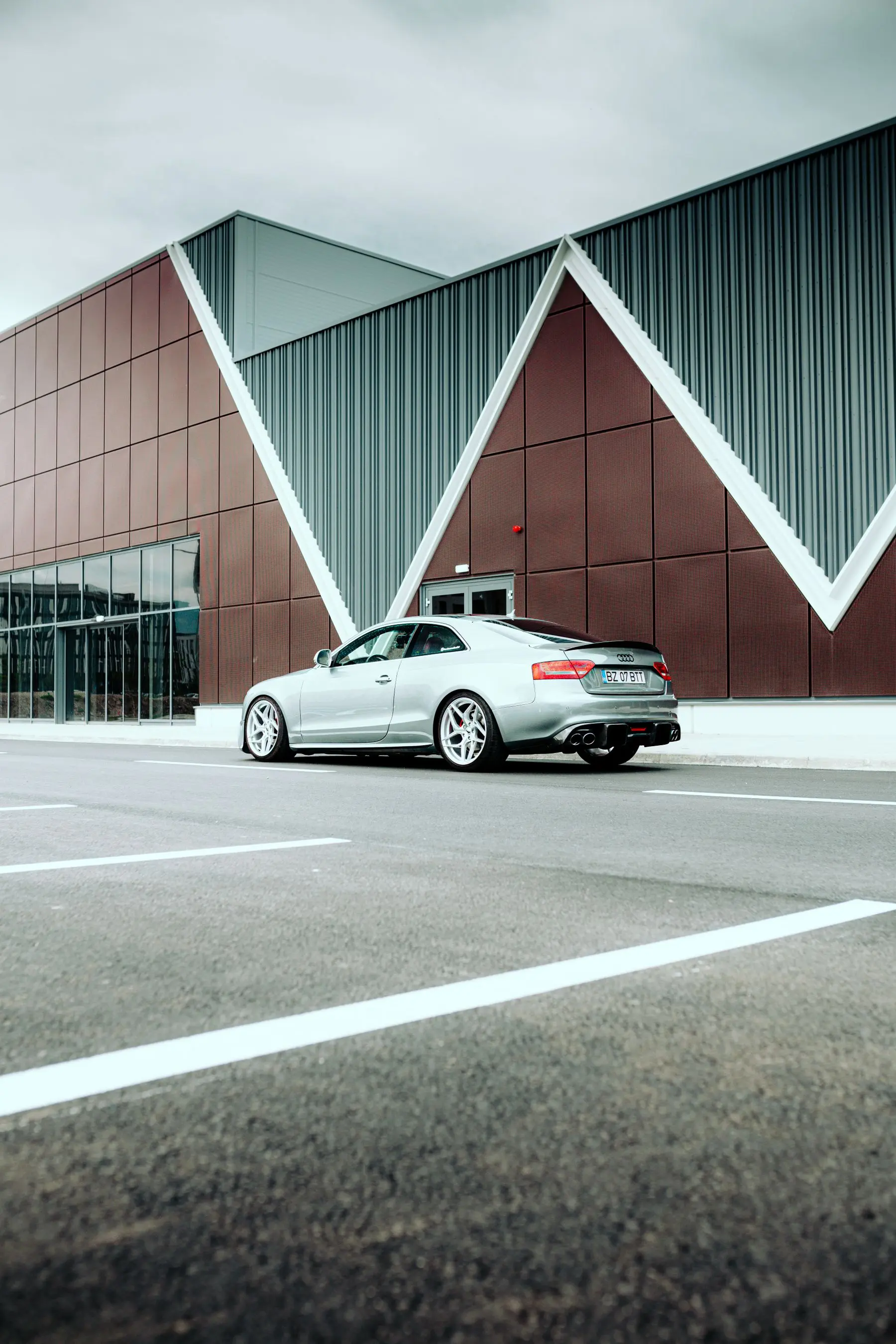 A silver coupe parked beneath the angular beams and geometric facade of a modern building.
