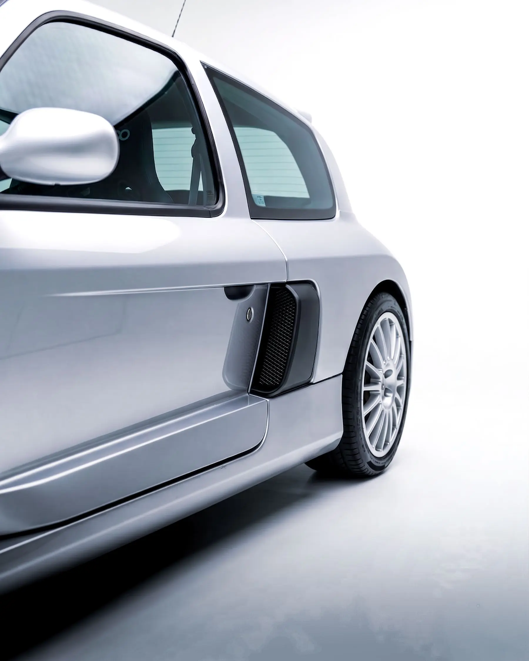 Close view of a silver sports car’s door, side intake, rear wheel, and curved roofline against a pale background.